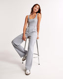 Woman wearing a light gray sleeveless jumpsuit sitting on a stool against a white background