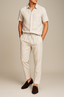 Man wearing a beige linen shirt and pants with brown shoes on a neutral background