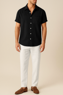 Man wearing a black short-sleeve shirt, white pants, and brown shoes on a beige background