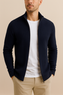 Man wearing a navy ribbed sweater over a white shirt and beige pants on a beige background