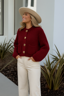 Woman wearing a red cardigan with gold buttons and a beige hat, standing outdoors.