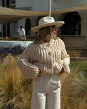 Woman wearing a beige knit sweater and wide-brimmed hat in an outdoor setting with plants and a building in the background.