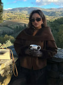 Woman in a brown coat and sunglasses holding a white mug with a scenic mountain background