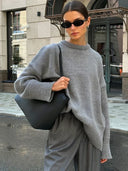 Woman wearing a gray sweater and sunglasses, holding a black bag on a city street.
