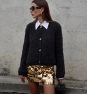 Woman wearing a black textured cardigan and gold sequin skirt against a textured wall.