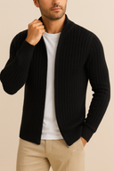 Man wearing a black ribbed sweater over a white shirt with beige pants on a beige background