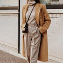 Person wearing a beige coat and pants with a black bag against a light-colored wall.