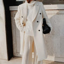 Person wearing a long white coat and white outfit with a black bag against a neutral wall.