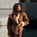 Woman wearing a brown coat with fur lining, standing against a textured wall.