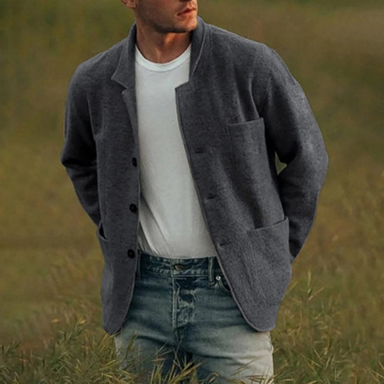 Man wearing a gray jacket over a white shirt and blue jeans in a grassy field.
