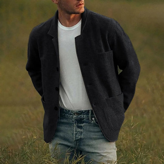 Man wearing a black jacket over a white shirt and blue jeans standing in a grassy field.