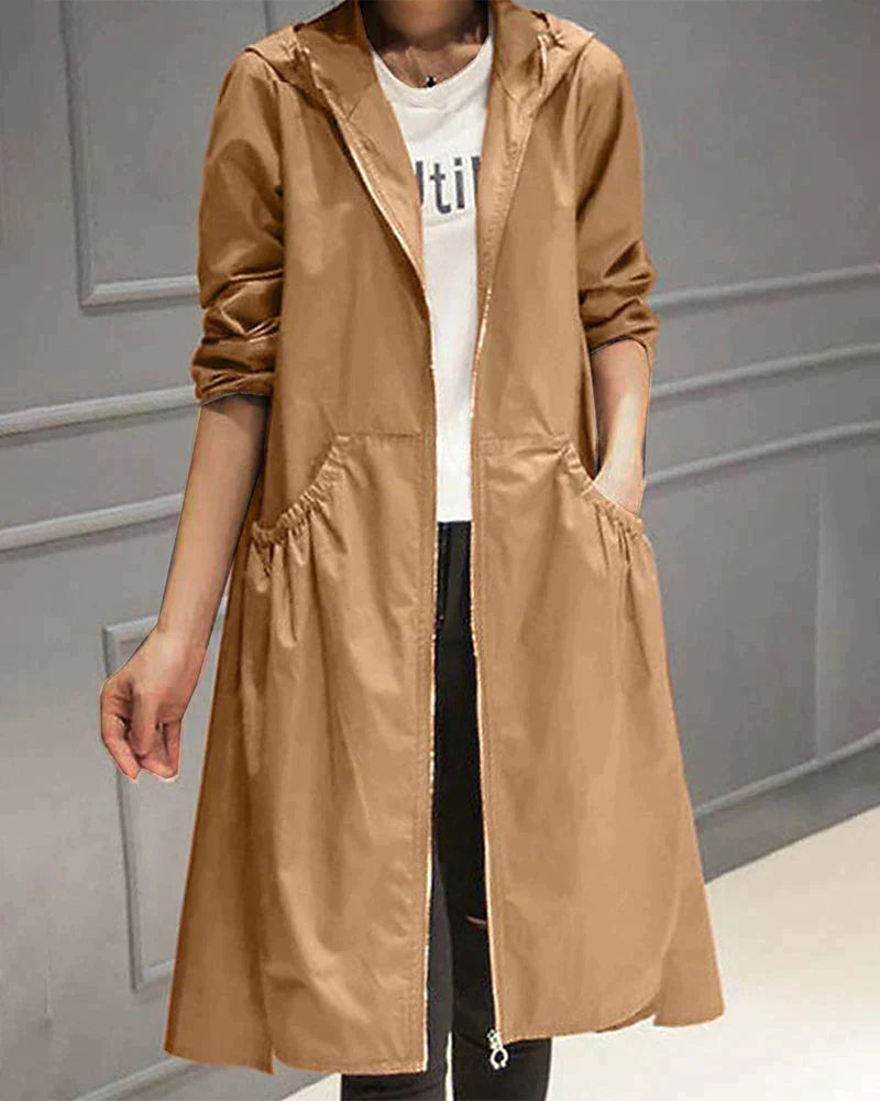 Person wearing a long beige coat over a white shirt with text, standing against a gray wall.