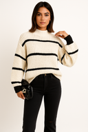 Woman wearing a cream sweater with black stripes and black pants on a plain background