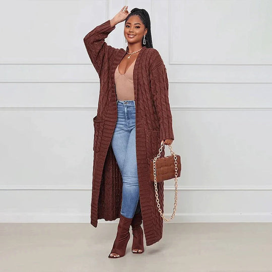 Woman wearing a long brown cardigan, blue jeans, and brown boots, holding a matching handbag against a white background.