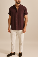 Man wearing a burgundy red short-sleeve shirt, white pants, and brown shoes on a beige background