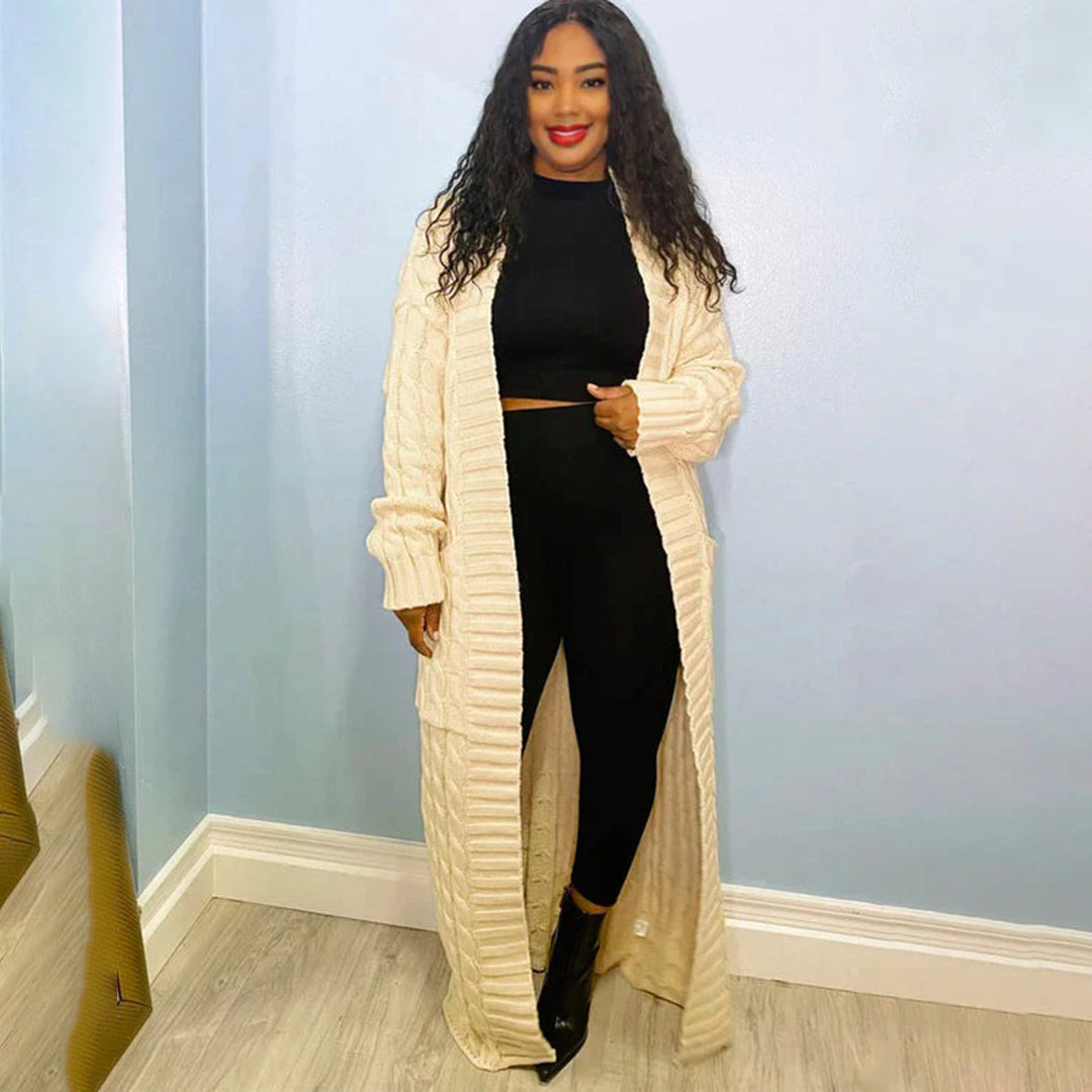 Woman wearing a long beige cardigan over a black outfit against a light blue wall.