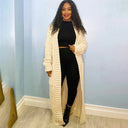 Woman wearing a long beige cardigan over a black outfit against a light blue wall.