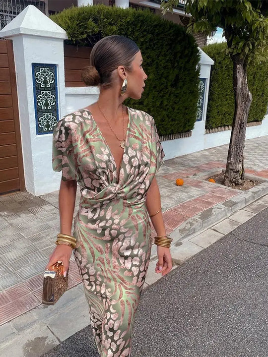 Woman in a patterned dress walking on a sidewalk with a white wall and greenery in the background.