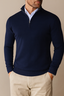 Man wearing a navy blue quarter-zip sweater over a light blue shirt with beige pants on a neutral background