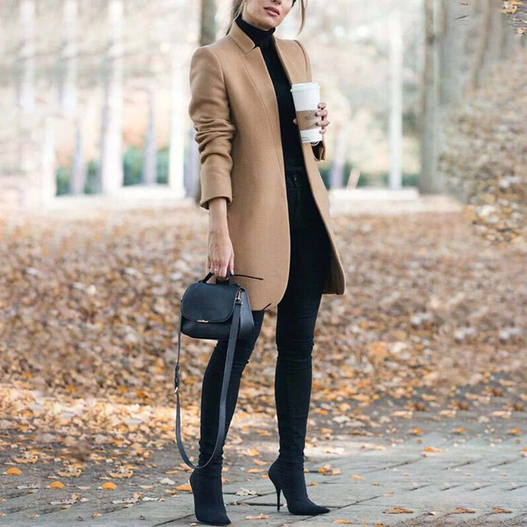 Woman in a tan coat holding a coffee cup, walking on a leaf-covered path.