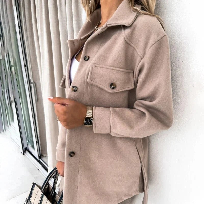 Person wearing a beige coat standing indoors with a neutral background