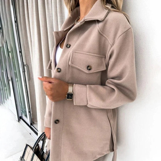 Person wearing a beige coat standing indoors with a neutral background