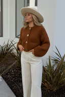 Woman wearing a brown knitted cardigan and white pants standing outdoors.