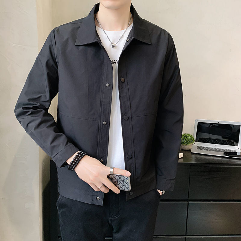 Person wearing a dark jacket over a white shirt, standing indoors with a laptop on a desk in the background.