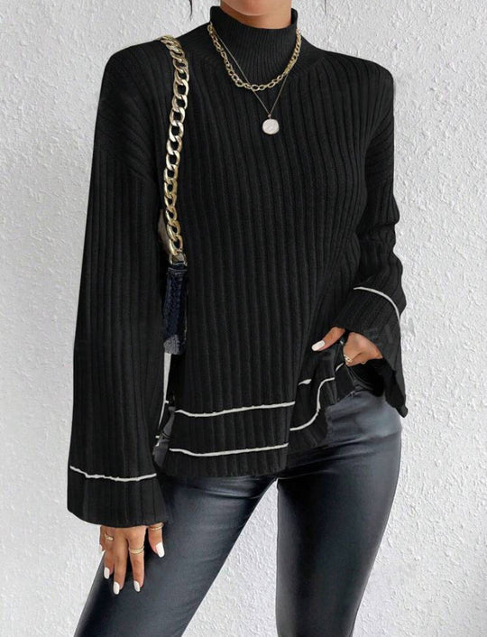 Person wearing a black ribbed sweater with white trim, black pants, and gold chain accessories against a light gray background.