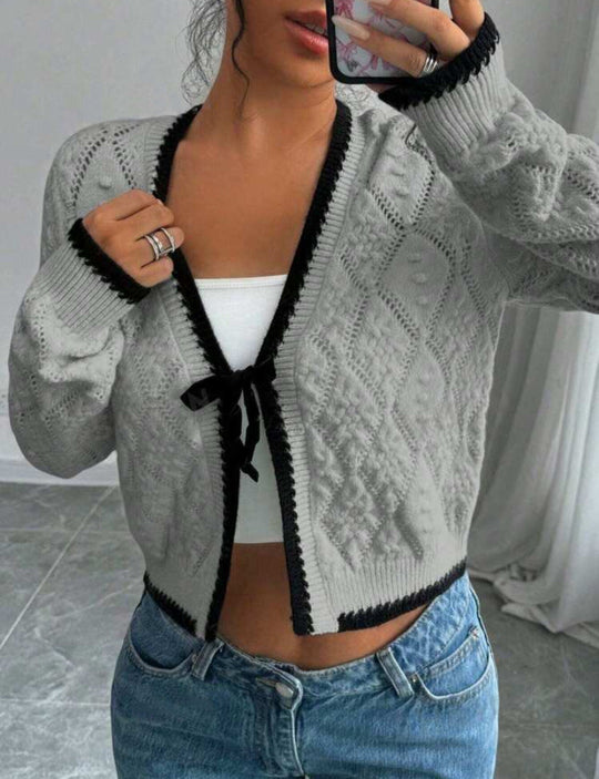 Person wearing a gray knitted cardigan with black trim, white top, and blue jeans indoors.
