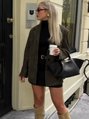 Woman in a green coat holding a coffee cup and black handbag, standing outside a building.