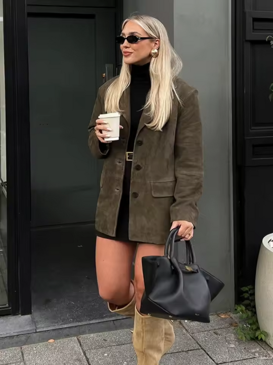 Woman in a brown coat holding a coffee cup and black handbag, standing against a dark door.