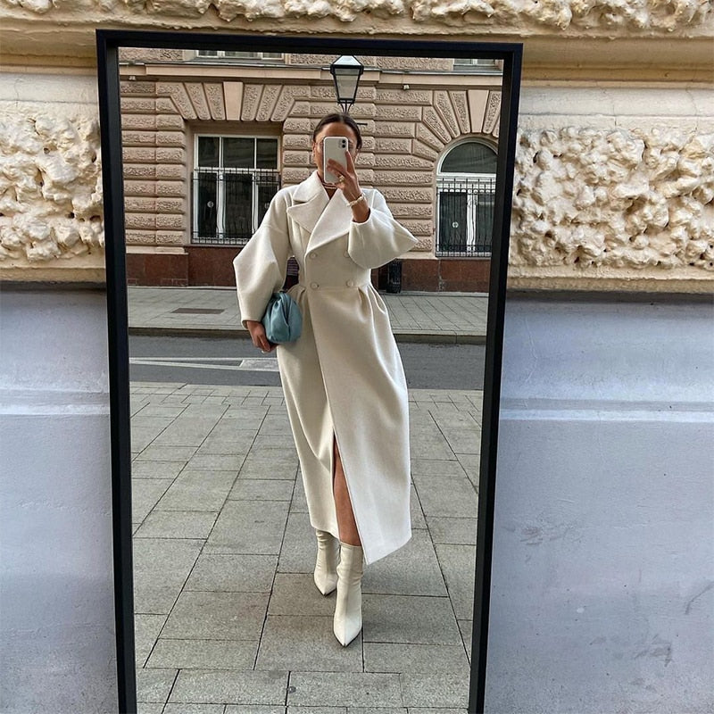 Person taking a mirror selfie wearing a long white coat and white boots in an urban setting.