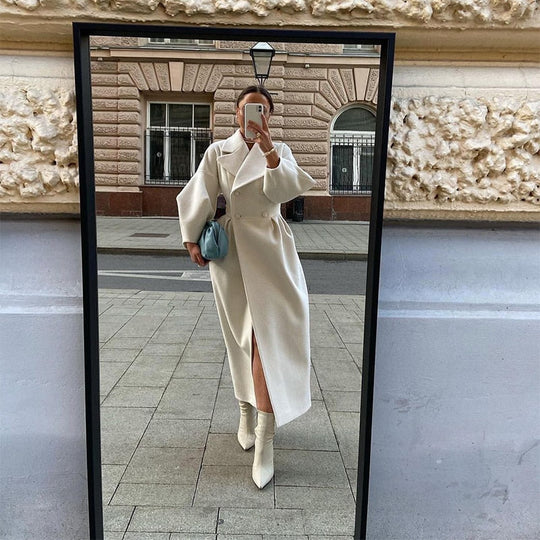 Person taking a mirror selfie wearing a long white coat and white boots in an urban setting.