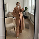 Person wearing a long beige coat taking a mirror selfie in a room with a couch and plant.