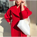 Woman in a red coat holding a white handbag, standing against a neutral background.