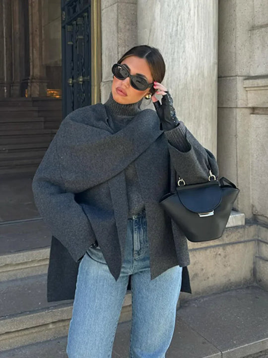 Person wearing a gray coat, blue jeans, and black gloves, holding a black handbag, standing against a stone building.