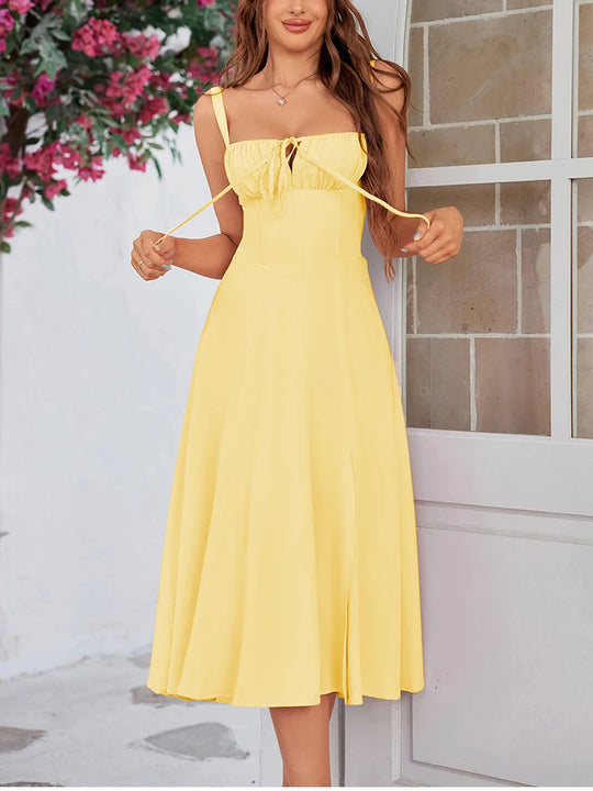 Woman wearing a yellow dress standing in front of a stone wall and plant.