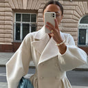 Person wearing a beige coat taking a selfie in front of a brick building.