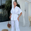Woman in a white outfit standing in a bright room with plants and furniture.