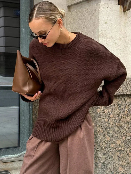 Woman wearing a brown sweater and holding a brown bag against a stone wall.