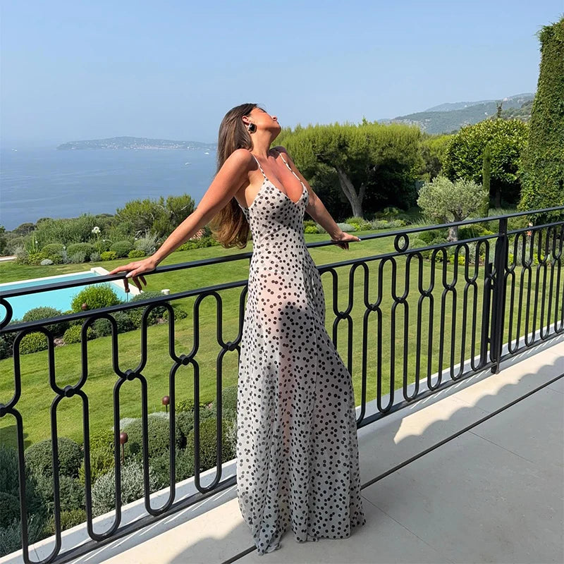 Woman in a polka dot dress standing on a balcony with a scenic view of the sea and greenery.
