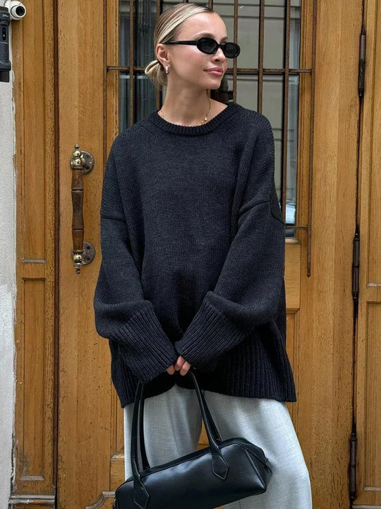 Person wearing a dark oversized sweater and sunglasses, holding a black handbag, standing in front of wooden doors.