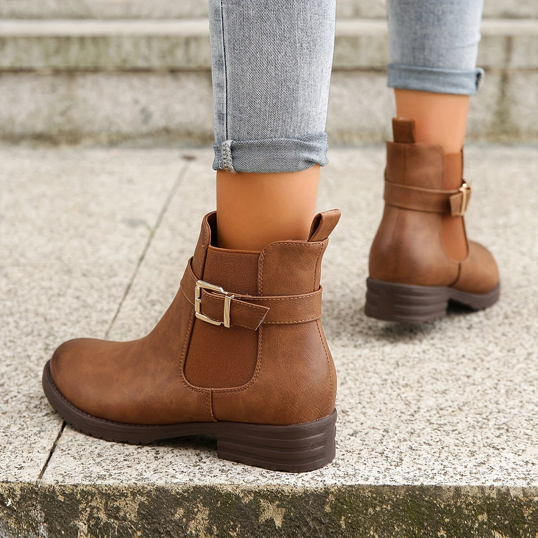 Brown ankle boots with buckles worn with rolled-up jeans on a concrete surface.