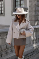 Woman in a stylish outfit with a white coat, beige hat, and shorts standing against a stone building.