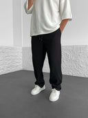 Person wearing a silver oversized shirt, black pants, and white sneakers in a minimalistic setting.