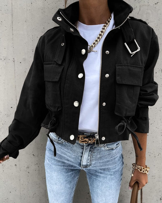 Person wearing a black jacket with white buttons, light blue jeans, and gold jewelry against a concrete wall.