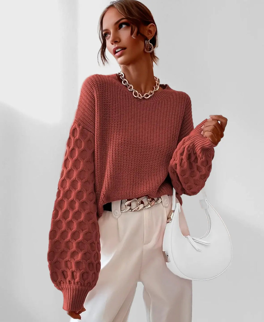 Woman wearing a rust-colored knitted sweater and white pants, holding a white handbag on a light background.