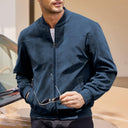 Man wearing a navy blue bomber jacket holding sunglasses, standing next to a car.